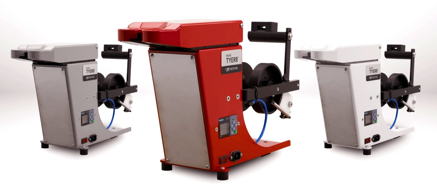 Three Bedford Mini Tyer machines, one red, one gray, one white, with spools and digital displays, on a light gray surface.