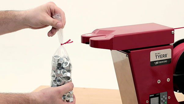 Hands tying a bag of nuts with red twist tie wire on a red "Mini Tyer" machine. Bedford branding visible.