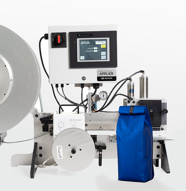 A silver table top machine that applies bag closures vertically with a blue coffee bag in front of the machine.