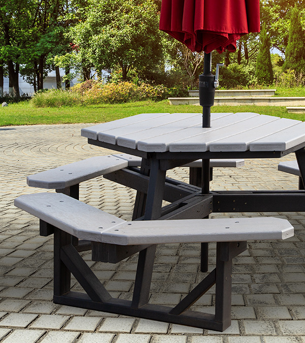 Hexagonal plastic lumber picnic table with gray top and black frame, red umbrella, on a stone patio in a park setting.