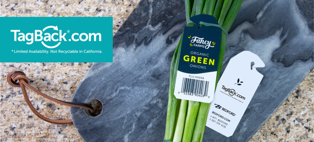 A close-up, angled shot of a bunch of organic green onions on a gray marble cutting board. The onions are tied with a dark blue tag that reads 'Fancy Farms Organic Green Onions' and includes a barcode and the PLU code '94068'. Next to the onions is a white tag with the 'TagBack.com' logo and the Bedford company information, including their website and phone numbers. In the upper left corner of the image is the 'TagBack.com' logo in a blue rectangle with the disclaimer '*Limited Availability. Not Recyclable in California.'