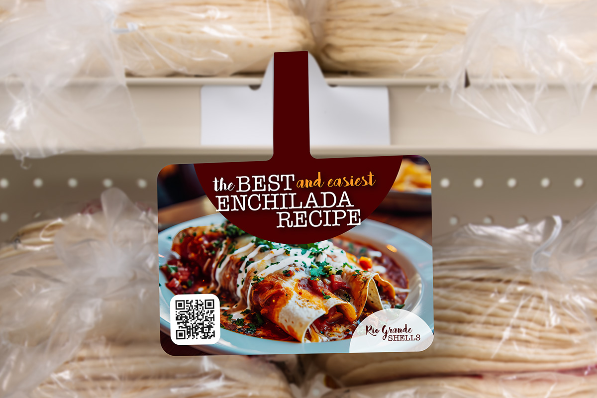 A shelf talker recipe card for "the BEST and easiest ENCHILADA RECIPE" hangs in a grocery store, with packages of tortillas visible behind it.
