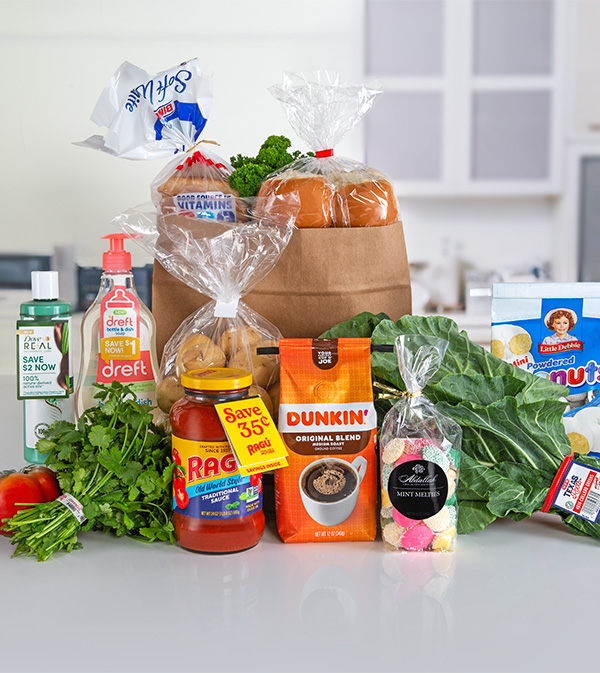 A still life photograph of various grocery items arranged on a light-colored countertop against a blurred kitchen background. The items are spread out and include: a white plastic bag with 'Soft White' branding floating in the upper left; a small paper bag containing baked goods wrapped in clear plastic, topped with a red tie; a clear plastic bag filled with dinner rolls in the center; a bottle of green liquid with 'REAL SAVE $2 NOW 100%' written on it; a bottle of 'dreft' dish soap; a jar of 'RAGU Old World Style TRADITIONAL SAUCE' with a 'Save 35¢' sticker; a bag of 'DUNKIN' ORIGINAL BLEND' coffee with a picture of a coffee cup; a clear bag of 'Abdallah MINT MELLETS' candies; a box of 'Little Debbie Powdered Mini Donuts'; a bunch of leafy greens; a red tomato; and a bunch of cilantro. The background shows a white kitchen counter with blurred cabinets and a window.