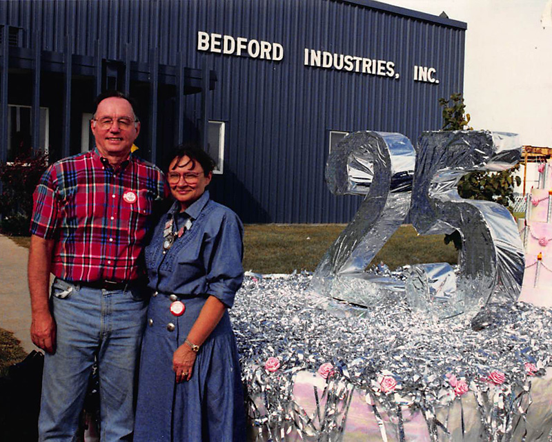 A man and woman stand in front of Bedford Industries manufacturing facility and a parade float decorated in silver tinsel with "25" as the centerpiece.