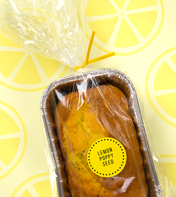 A yellow twist tie secures a plastic overwrap on a lemon poppy seed loaf of bread in a tin bread pan.