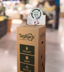 A tall cardboard box with small deposit hole, TagBack logo, and instructions: “Recycle Tags & Ties Here”