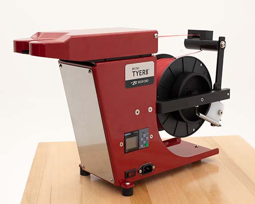 Close-up of the Bedford Mini Tyer Pro twist tie machine in a ruby red finish, placed on a wooden surface. The machine features a spool holder, control panel, and sleek, compact design, suitable for efficient twist tying in packaging applications.