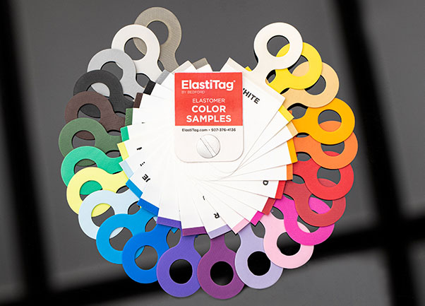 A fan-shaped display of ElastiTag color samples. The samples are arranged in a rainbow pattern, with each color represented by a small, circular tag. The tags are attached to a white card with the ElastiTag logo. The colors shown include white, yellow, orange, red, pink, purple, blue, green, and black.