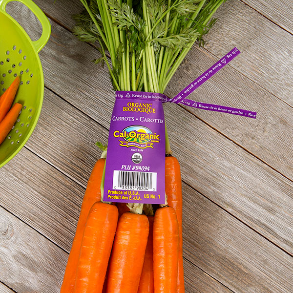 A bunch of fresh, orange carrots with green tops, tied together with a purple produce label. The label indicates that they are organic and produced in the USA.