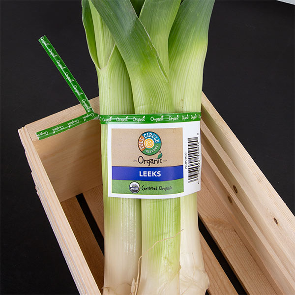 A bunch of fresh, green leeks tied together with a produce twist tie label that says "Full Circle Organic Leeks" certified organic. The leeks are in a wooden crate.