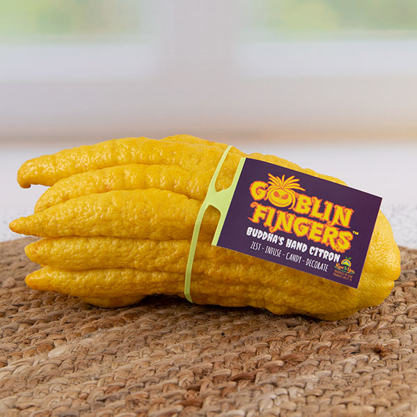 A Buddha’s hand citron labeled "Goblin Fingers" with a branded ElastiTag. The bright yellow fruit has elongated, finger-like segments and sits on a woven mat. The tag highlights the product’s playful branding with the text “Goblin Fingers” in bold, colorful letters, along with “Buddha's Hand Citron” and suggestions for uses: zesting, infusing, candying, and decorating.