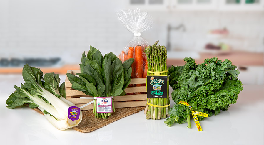 An assortment of fresh produce displayed on a kitchen counter with various tags and ties. Items include bok choy with a purple ElastiTag, spinach bunch with a labeled PushTag, bagged carrots in a wooden crate, asparagus tied with a yellow band featuring branding, and kale with a yellow ElastiTag. The tags highlight different labeling and branding options for produce packaging, with each type of tag suited to the specific produce.