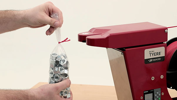 A person is holding a bag of nuts in their left hand and is about to place it into a red bag tying machine. The machine has a spool of red plastic ties.