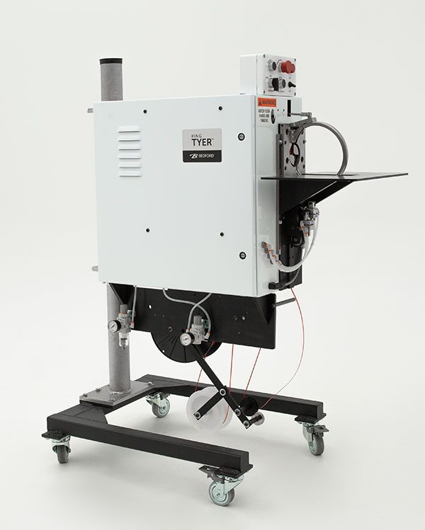 A white industrial bag tying machine with a black base and wheels. It has several levers and controls, and a spool of red plastic twist ties.