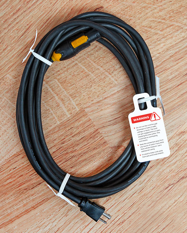 A coiled black extension cord with a warning Ring Tag label attached to the white twist tie. The label has a red exclamation point and lists safety precautions.