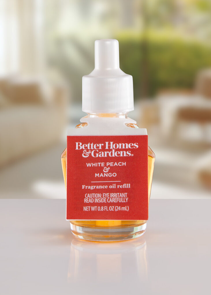 Close-up photo of a Better Homes & Gardens fragrance oil refill bottle. The ElastiTag label is red with white text and features the brand logo and scent name. The bottle contains a yellow liquid.