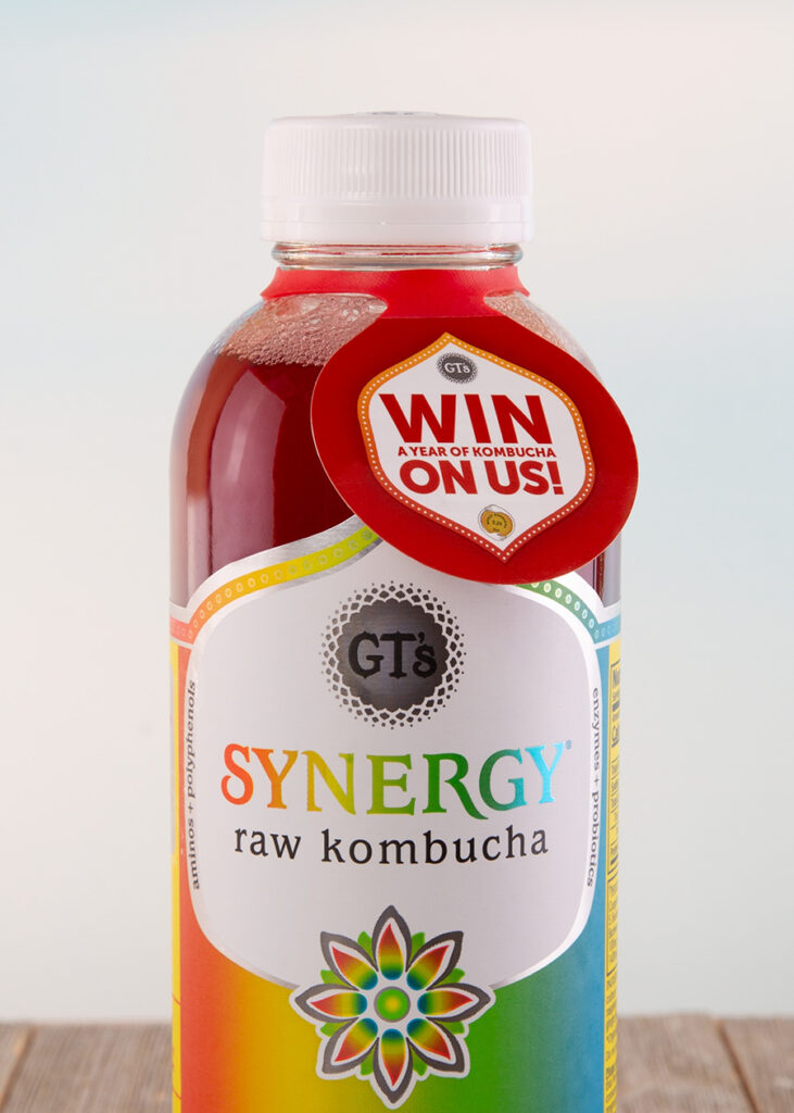 Close-up photo of a GT's Synergy Raw Kombucha bottle with a "Win a Year of Kombucha" ElastiTag hang tag. The bottle shows a colorful gradient design and details about the kombucha's ingredients and benefits.