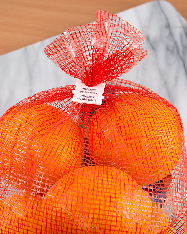 Close-up of a red mesh bag filled with oranges, with a white bag closure label that says "Product of Mexico."